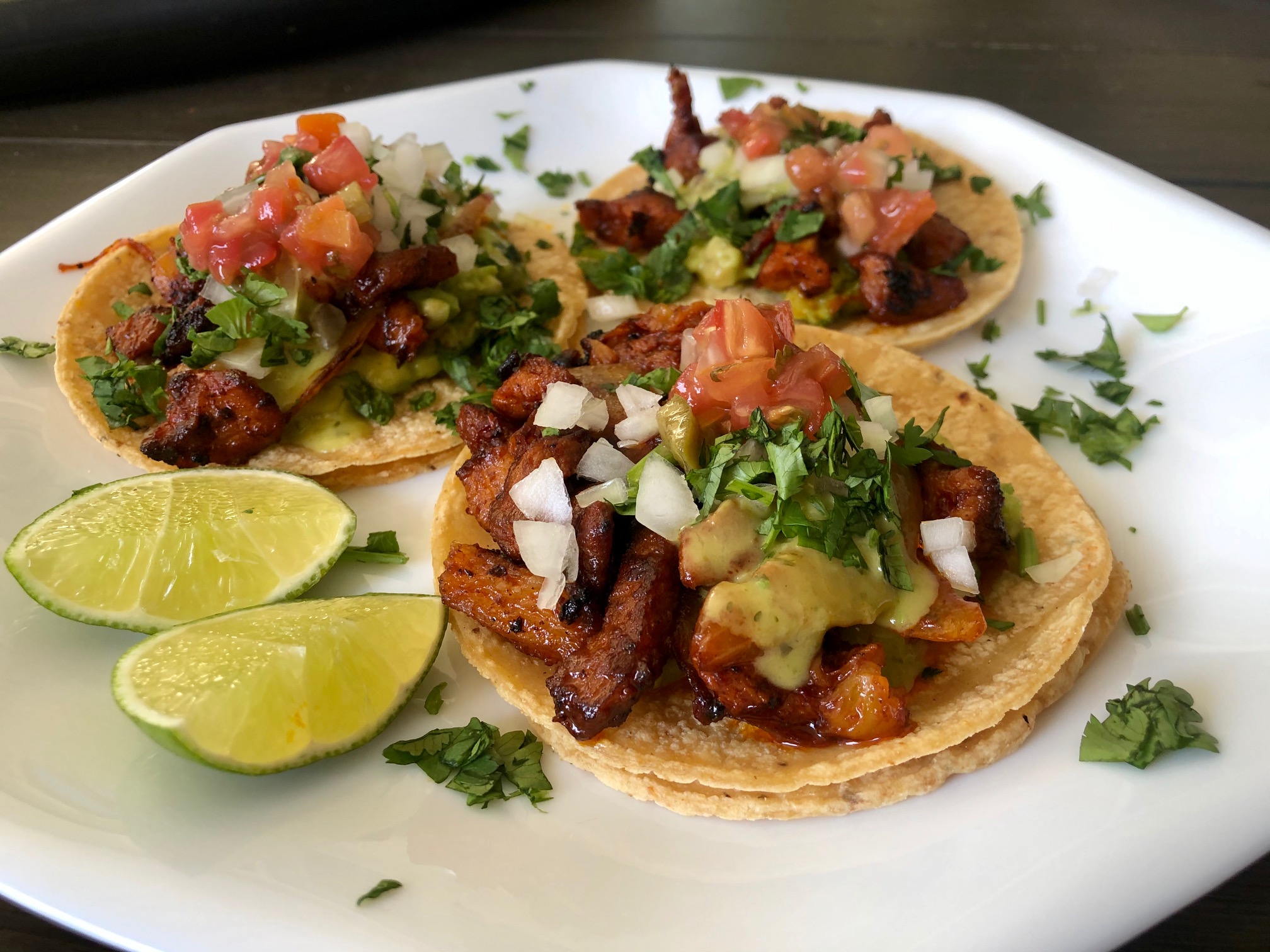 A platter of tacos al pastor on a warm-toned kitchen table, topped with chopped onion, cilantro, and pineapple, with lime wedges on the side