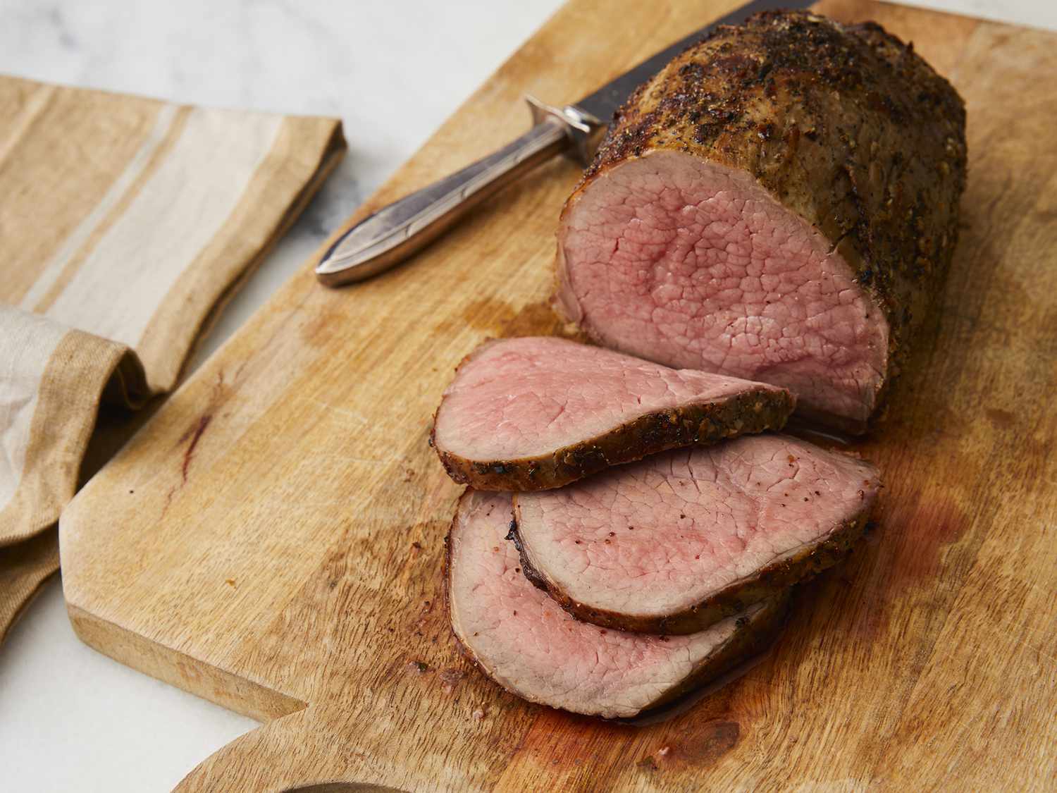 A platter of thinly sliced eye of round roast with a garlic herb crust, resting juices on the cutting board, and a small bowl of pan gravy on a cozy dining table