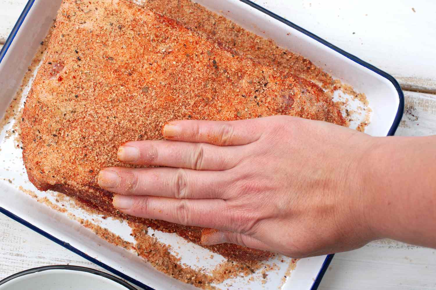 A pork shoulder on a cutting board being rubbed with salt and liquid smoke by a cook's hands on a clean kitchen counter