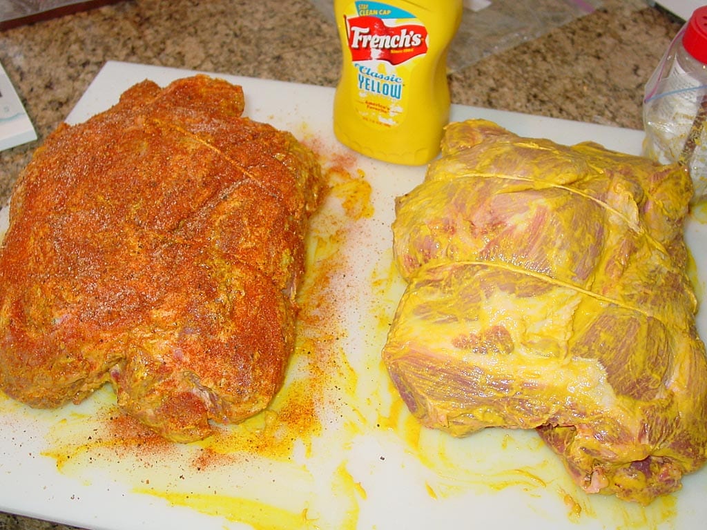 A pork shoulder on a tray being rubbed with a thin layer of yellow mustard by a cook's hands in a home kitchen