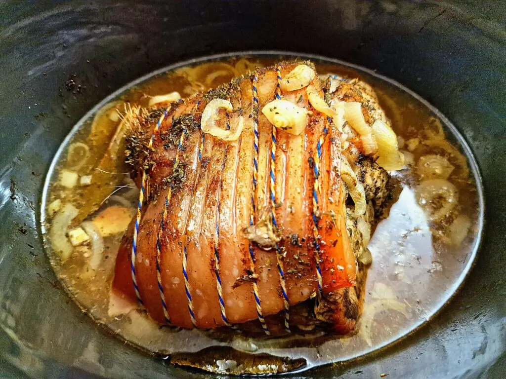 A pork shoulder sitting in a slow cooker as BBQ sauce is being poured over it, with onions visible around the edges