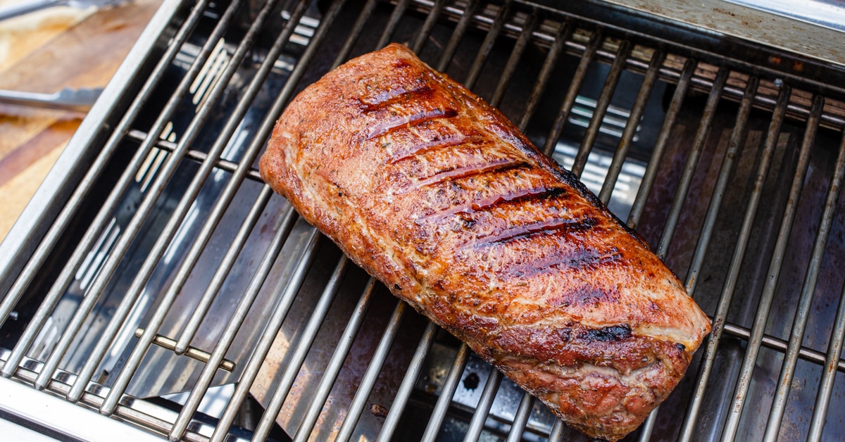 A pork tenderloin being turned with tongs on hot grill grates with visible grill marks and light smoke rising