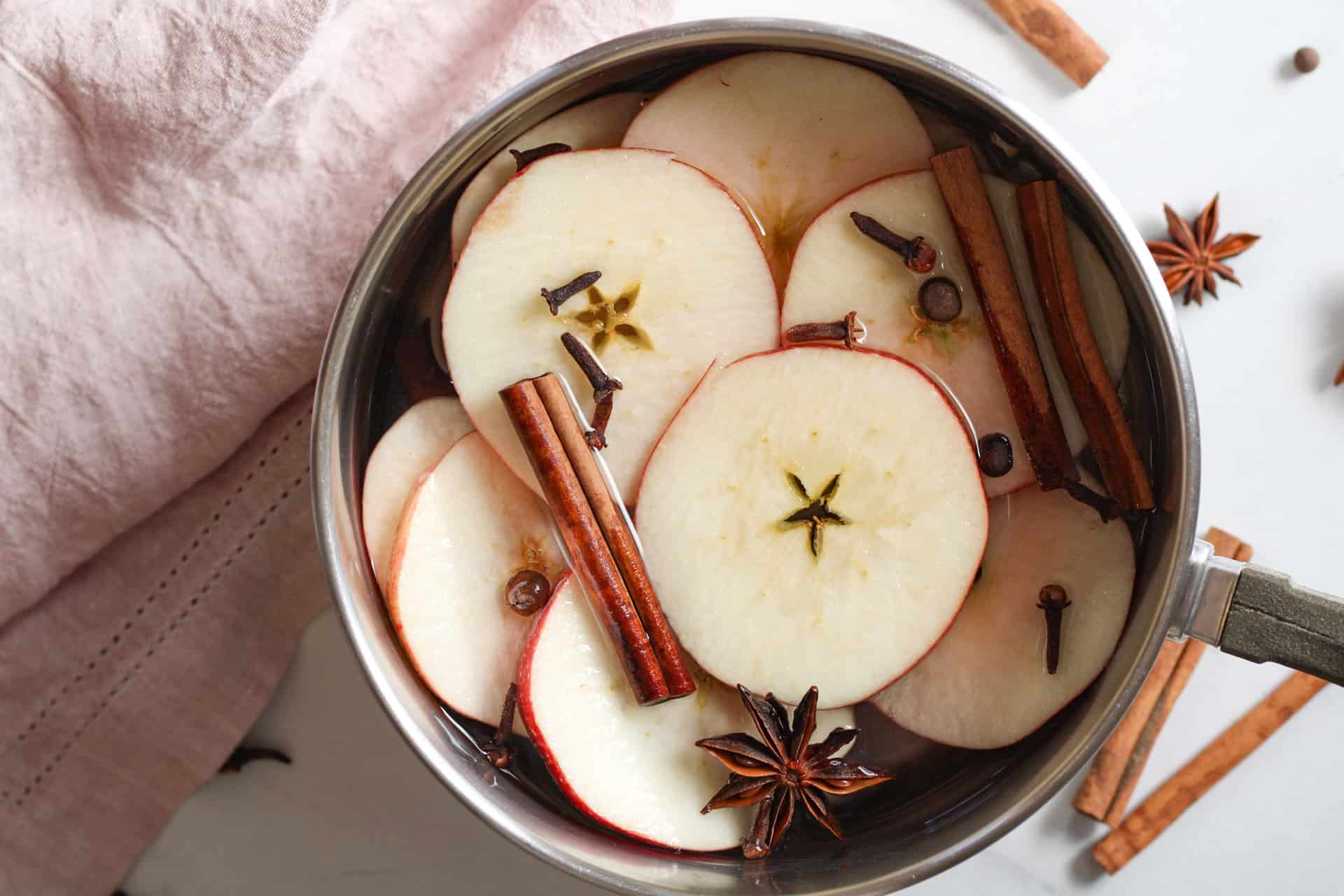 A pot of apple cider gently simmering with cinnamon sticks and orange peel floating on the surface on a home stove
