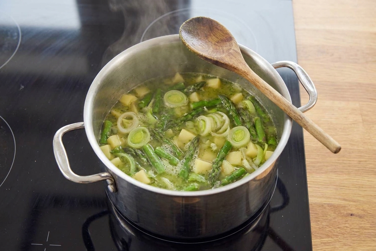 A pot of asparagus, leeks, and broth gently simmering on a stovetop with a wooden spoon resting on the rim