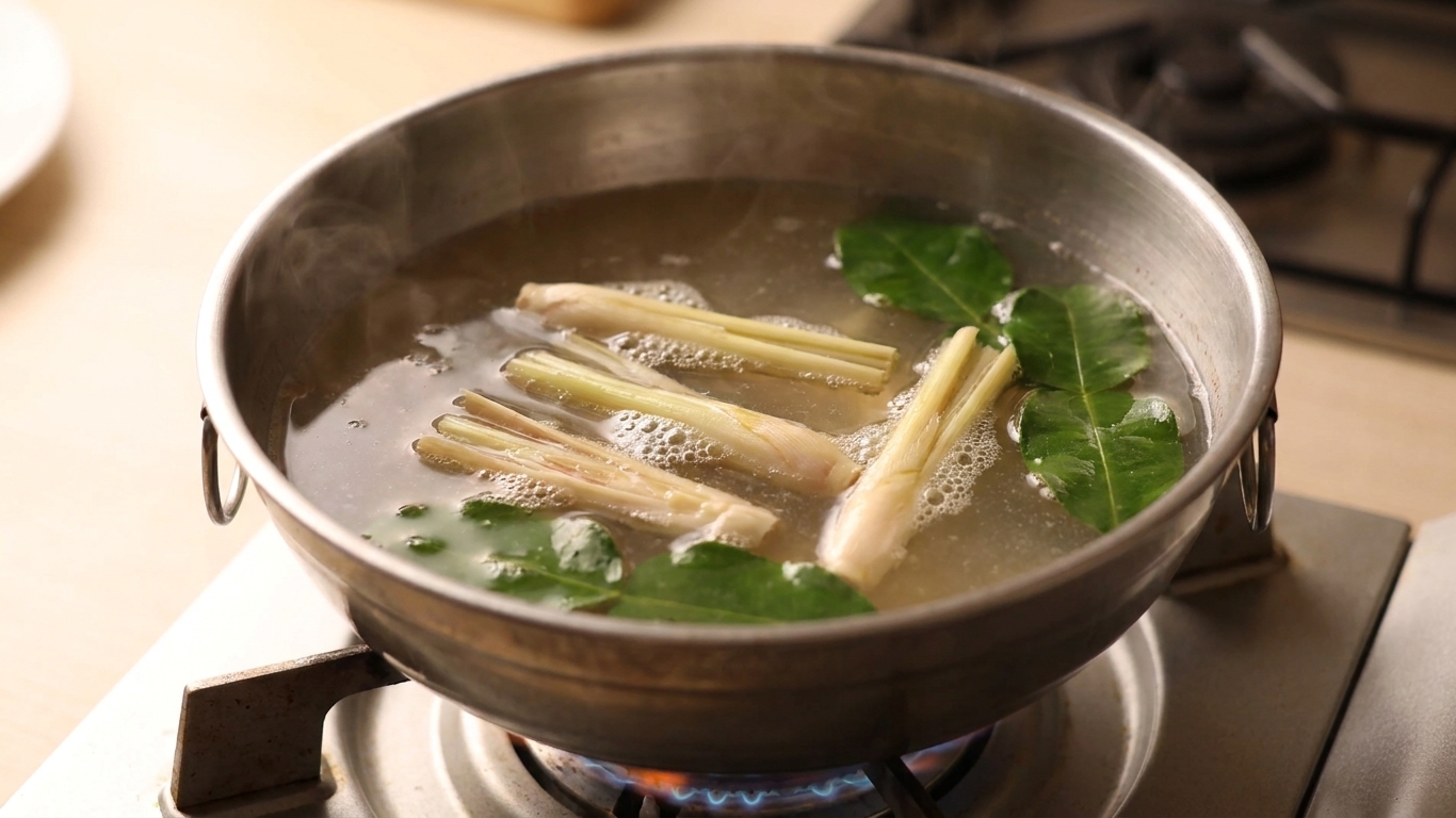 A pot of broth starting to heat on a stovetop with lemongrass pieces and lime leaves floating on the surface
