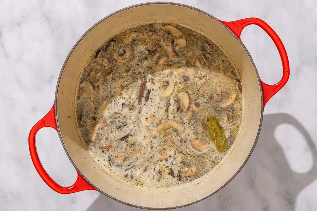A pot of creamy mushroom soup simmering on a stovetop with visible slices of mushrooms and a wooden spoon resting on the rim, warm kitchen lighting