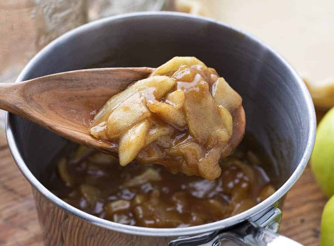 A pot of glossy apple pie filling with tender apple slices and cinnamon, simmering on a stovetop in a cozy home kitchen