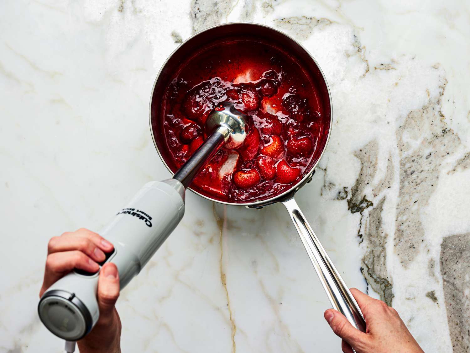 A pot of strawberry sauce being blended with an immersion blender on a stovetop, with a towel on the counter nearby