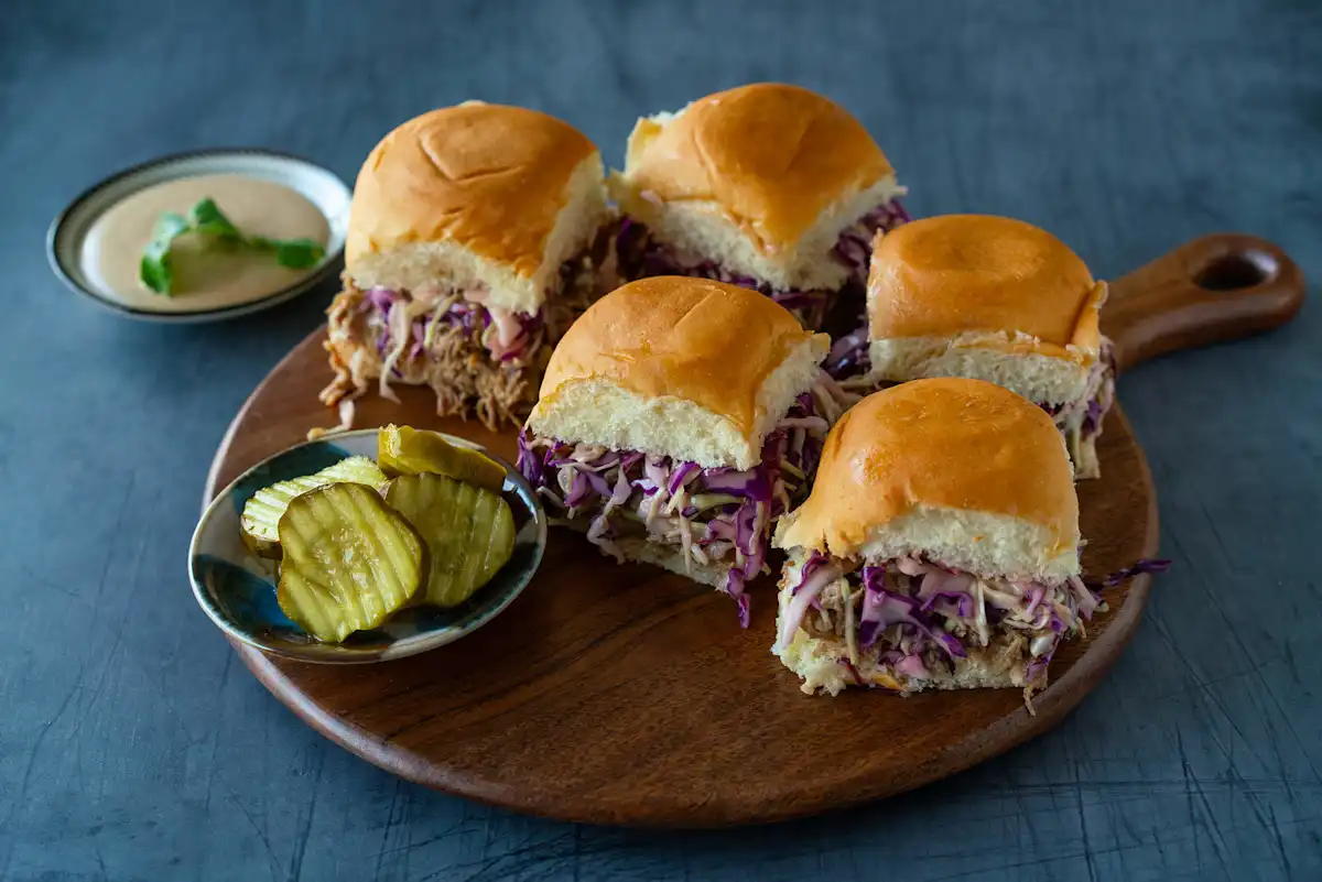 A pulled pork sandwich piled high with juicy shredded pork, tangy slaw, and a glossy vinegar finishing sauce on a toasted bun, set on a wooden board on a bright kitchen table
