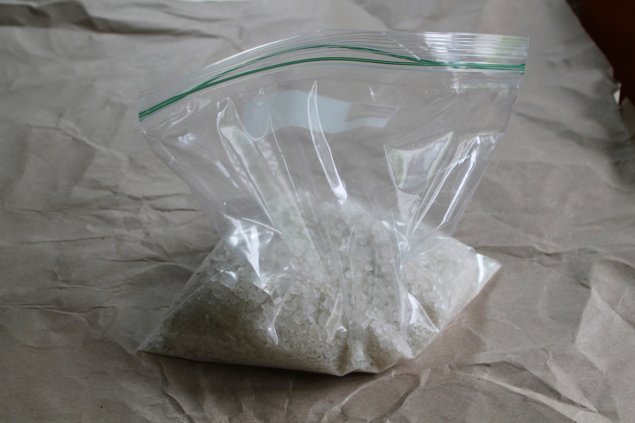 A quart-size and gallon-size resealable bag next to a bowl of ice cubes and a dish of coarse rock salt on a kitchen counter