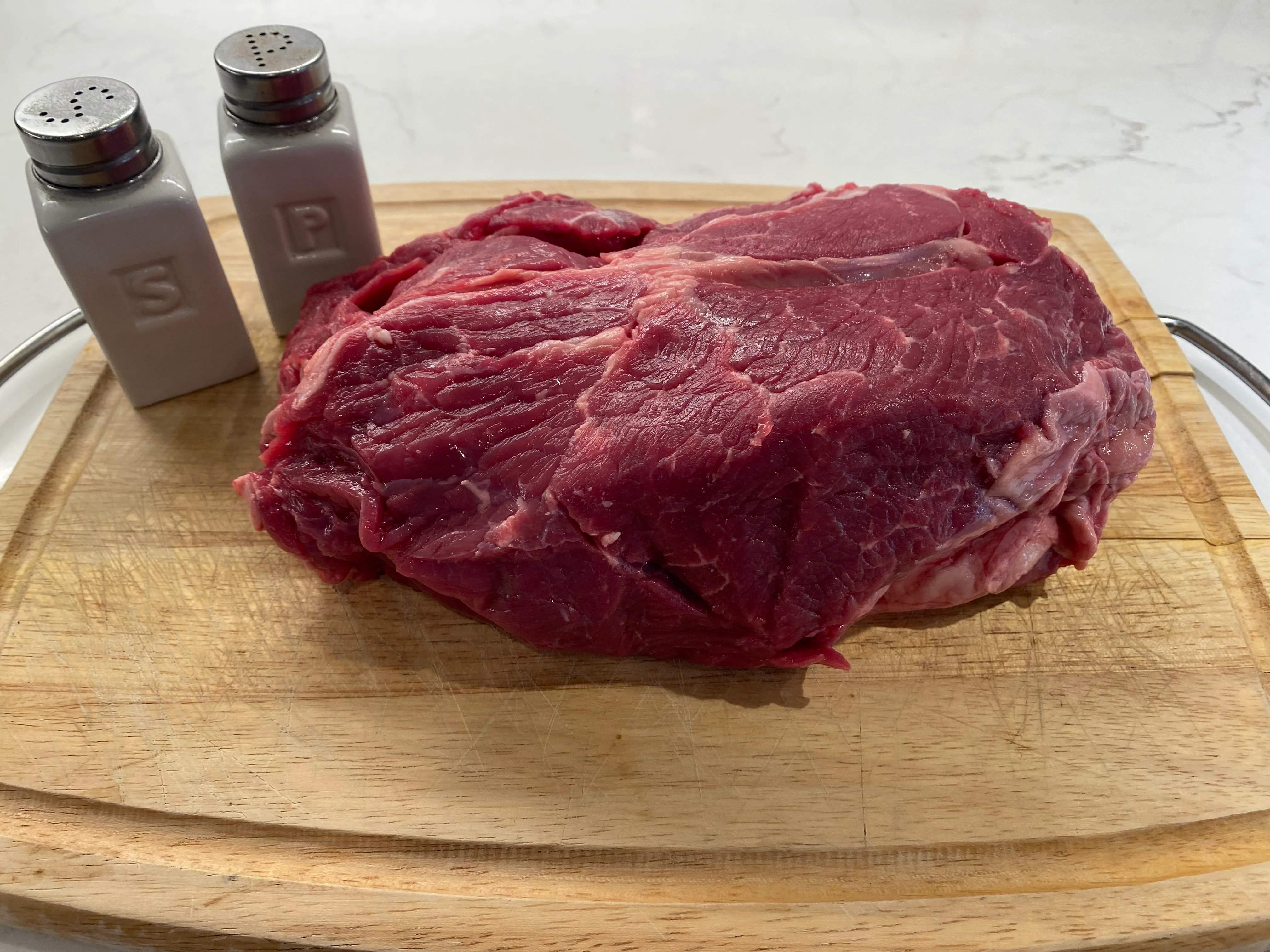 A raw chuck roast on a wooden cutting board being seasoned with salt and pepper next to chopped onions and carrots