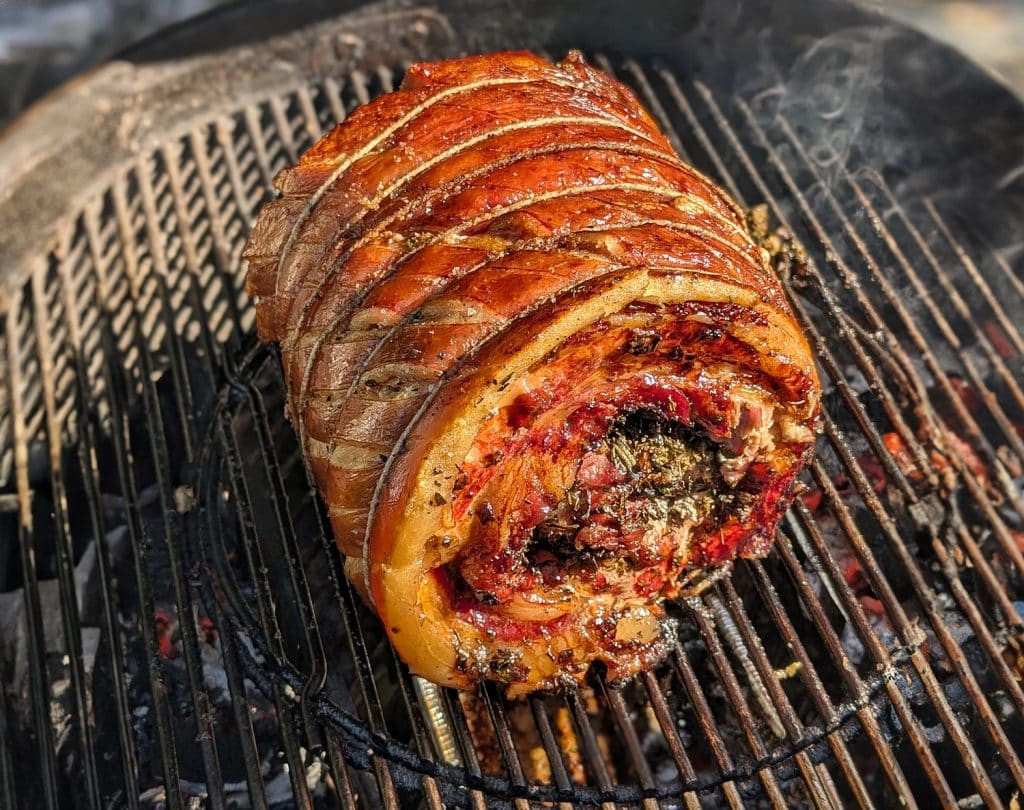 A raw pork belly rolled into a tight log and tied with kitchen twine on a wooden cutting board with garlic, lemongrass, and bay leaves nearby