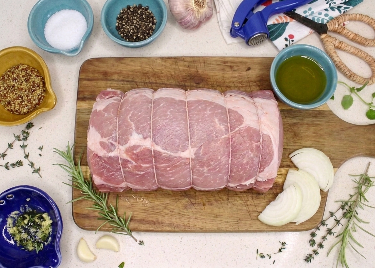 A raw pork roast on a cutting board surrounded by small bowls of salt, pepper, garlic, mustard, olive oil, rosemary, thyme, and onions on a bright kitchen counter