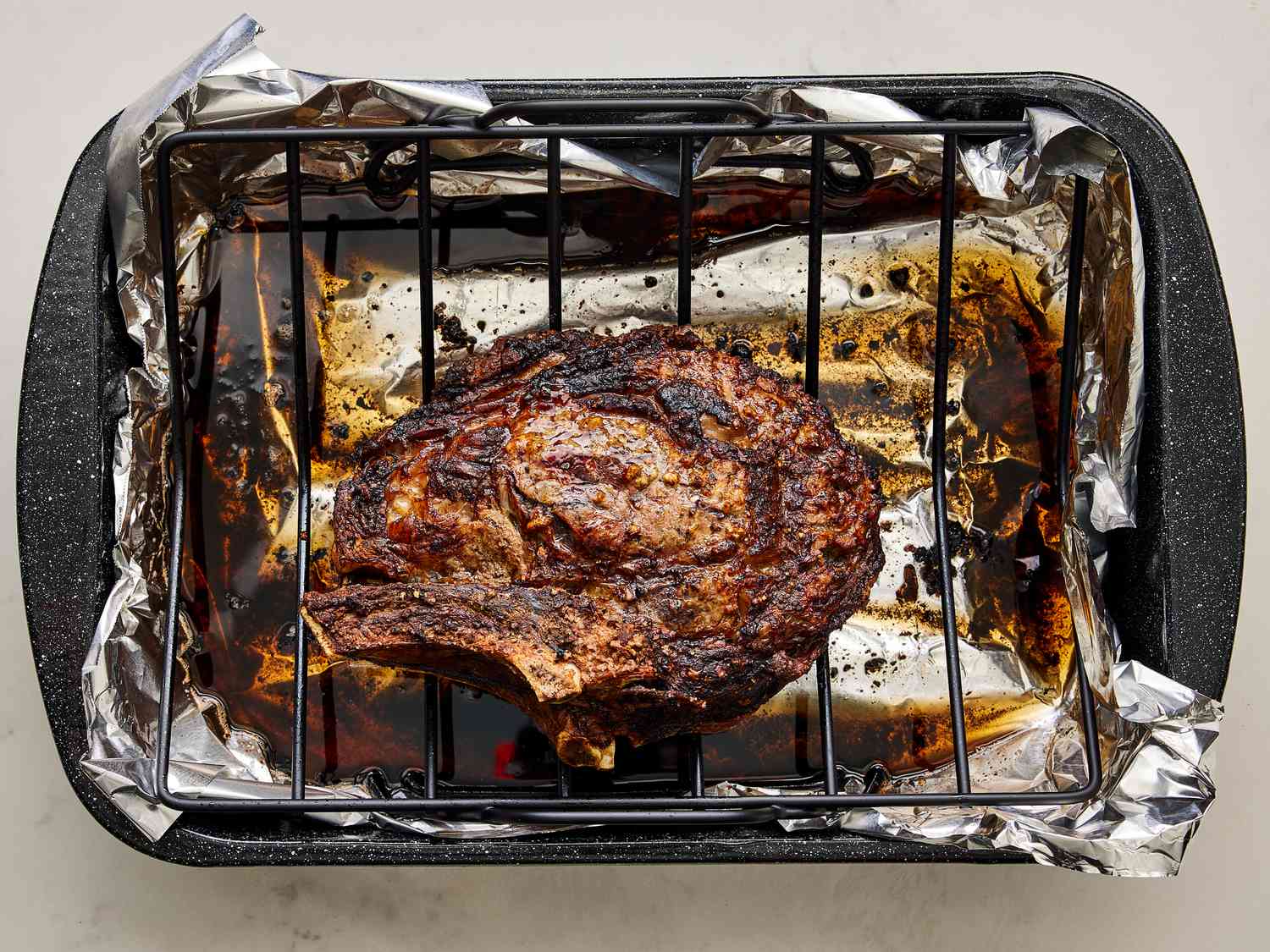 A raw prime rib roast seasoned with salt, pepper, and herbs on a rimmed baking sheet, with small bowls of garlic and rosemary nearby on a clean kitchen counter