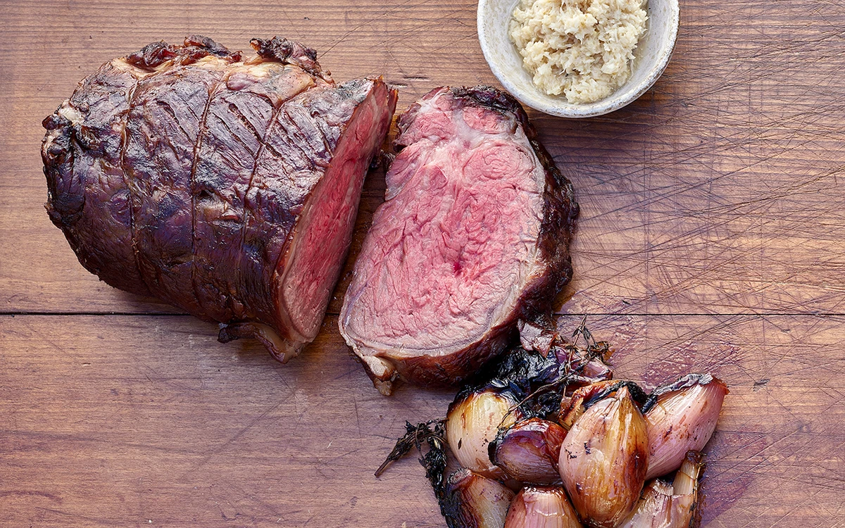 A raw ribeye roast on a rimmed sheet pan surrounded by small bowls of salt, pepper, minced garlic, chopped rosemary, thyme, olive oil, and Dijon mustard on a bright kitchen counter
