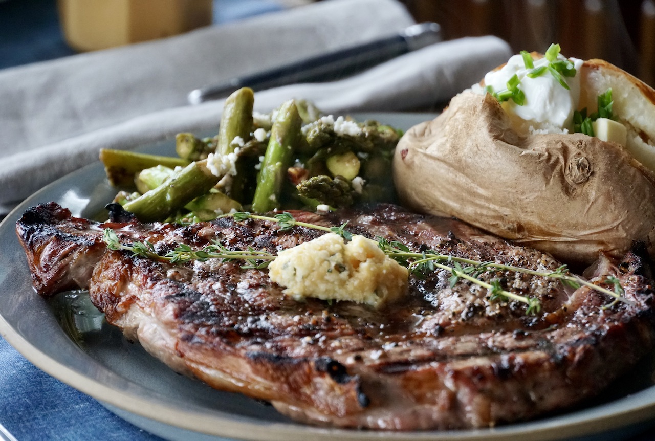 A raw ribeye steak on a plate being patted dry with paper towels next to small bowls of kosher salt, black pepper, and minced garlic on a home kitchen counter