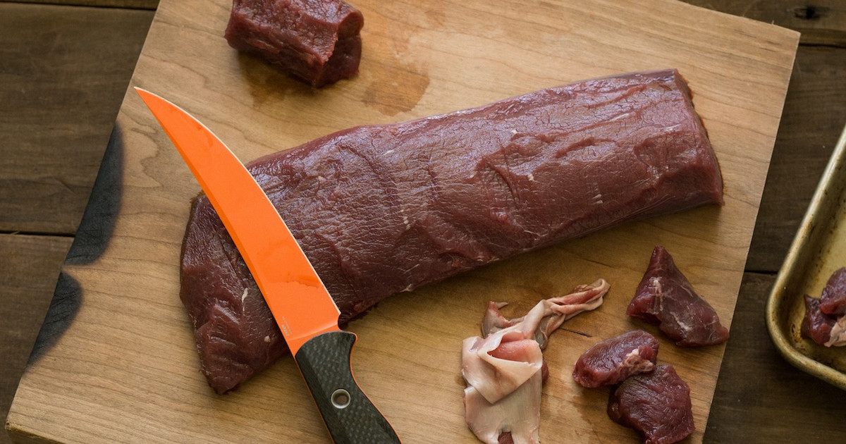 A raw venison backstrap trimmed of silver skin on a wooden cutting board with a small pile of salt and pepper nearby