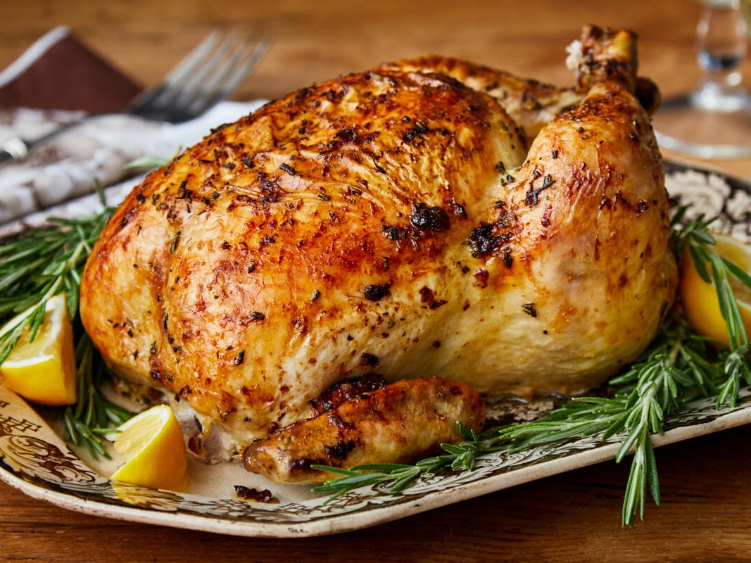 A raw whole chicken on a rimmed baking sheet with small bowls of kosher salt, black pepper, paprika, olive oil, butter, lemon, and fresh thyme arranged nearby