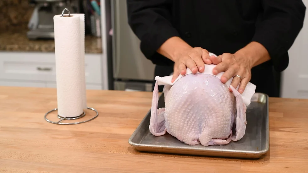 A raw whole turkey being patted dry with paper towels on a roasting rack set over a sheet pan on a clean kitchen counter