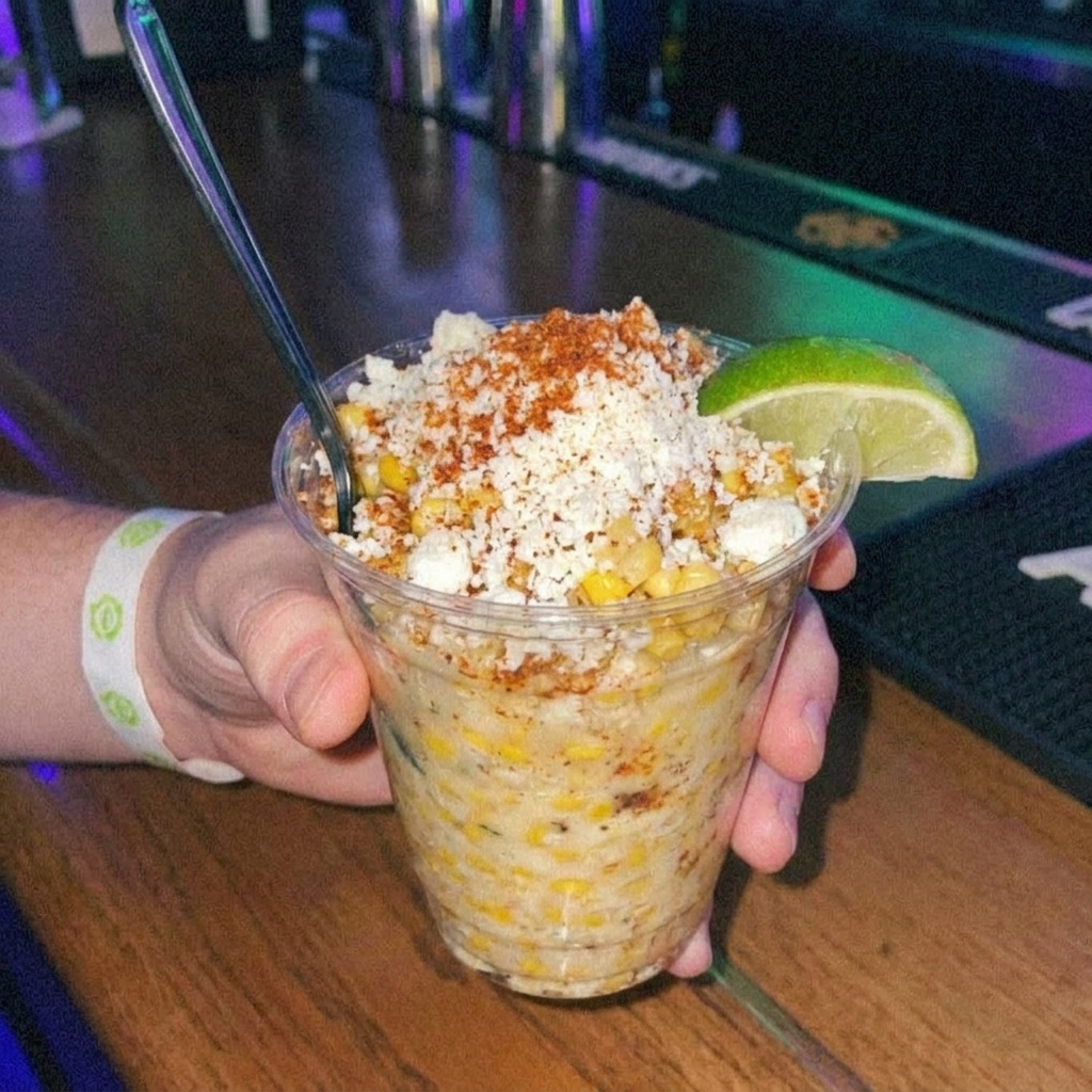 A real close-up photo of a cup of esquites topped with crumbled cotija and chili powder, with a lime wedge on the rim and a spoon tucked inside