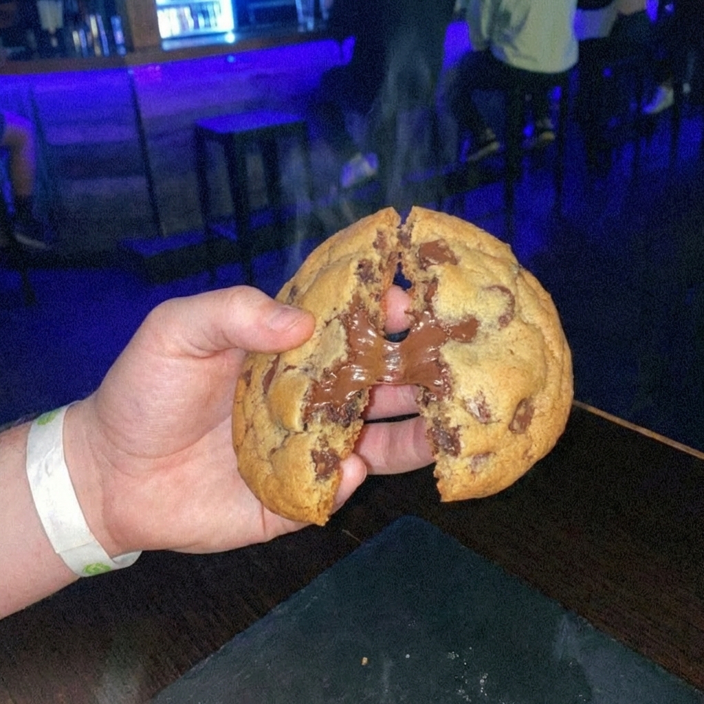 A real photo of a hand breaking a warm cookie in half showing a gooey chocolatey soft center