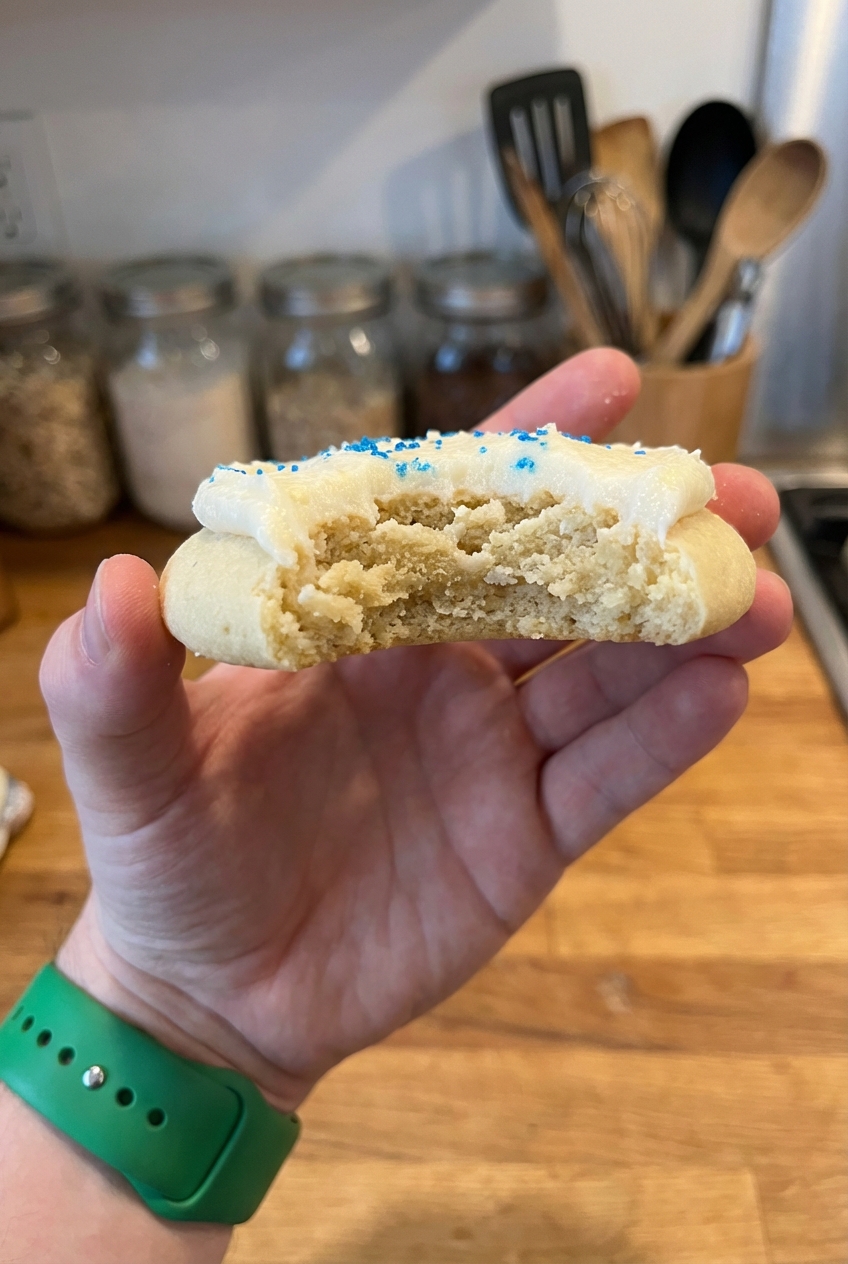 A real photo of a hand holding a thick frosted cookie with a bite taken out showing a soft chewy interior