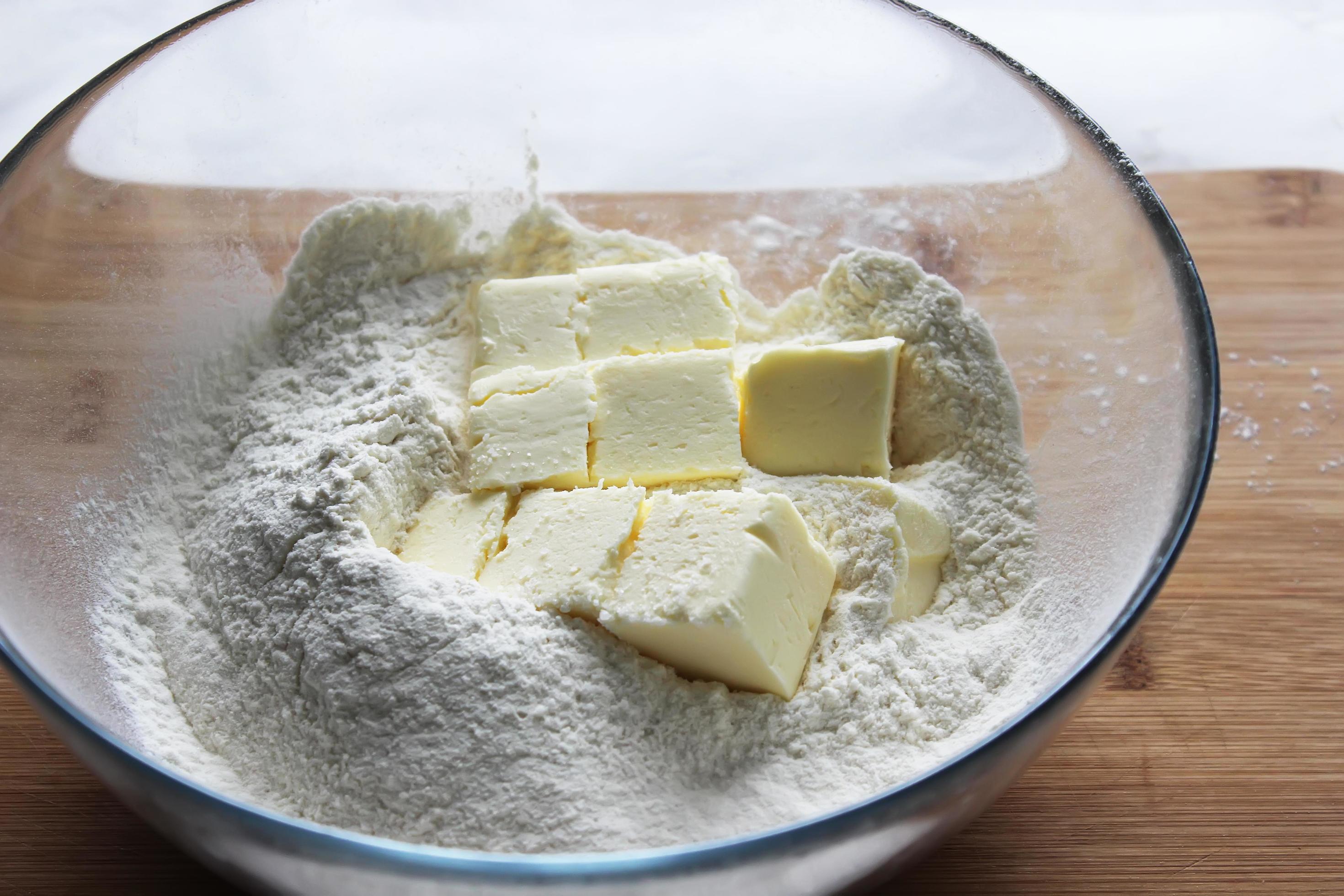 A real photo of cold butter cubes sitting in a bowl of flour with a pastry cutter nearby on a kitchen counter