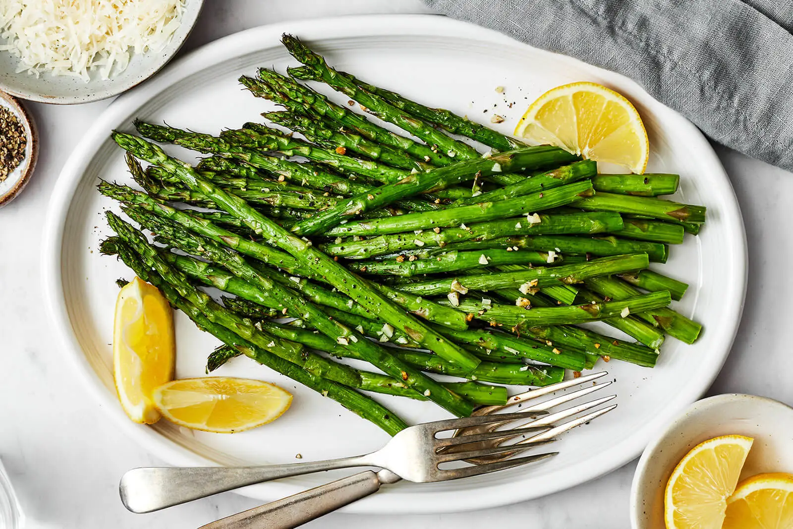 A real photo of crispy-tender air fryer asparagus on a white plate with lemon wedges and visible flecks of garlic and black pepper, warm kitchen lighting