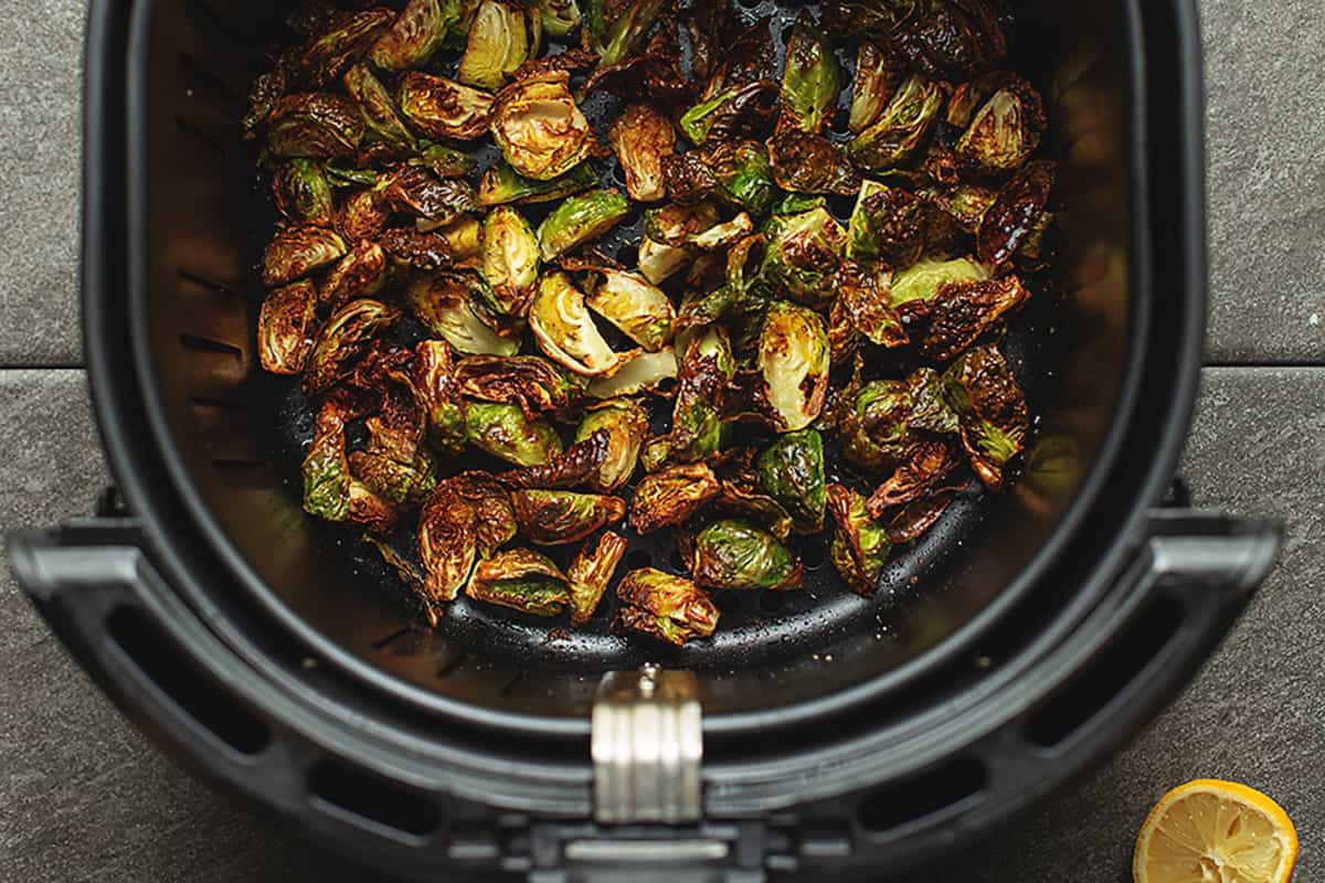 A real photograph of Brussels sprouts roasting in an air fryer basket with browned, crispy outer leaves and a light sheen of oil