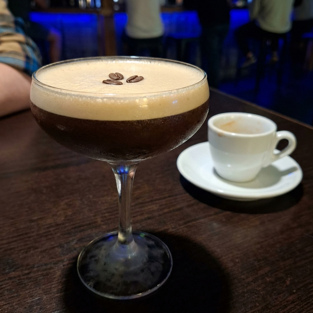 A real photograph of a chilled espresso martini in a coupe glass with a thick creamy foam layer and three coffee beans on top, on a dark bar surface with a small espresso cup in the background