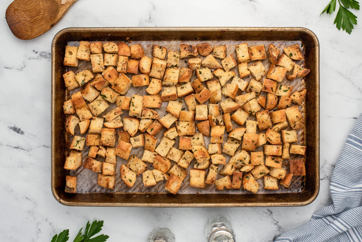 A real photograph of bread cubes tossed with olive oil on a sheet pan, ready to bake into croutons on a home kitchen counter in natural window light