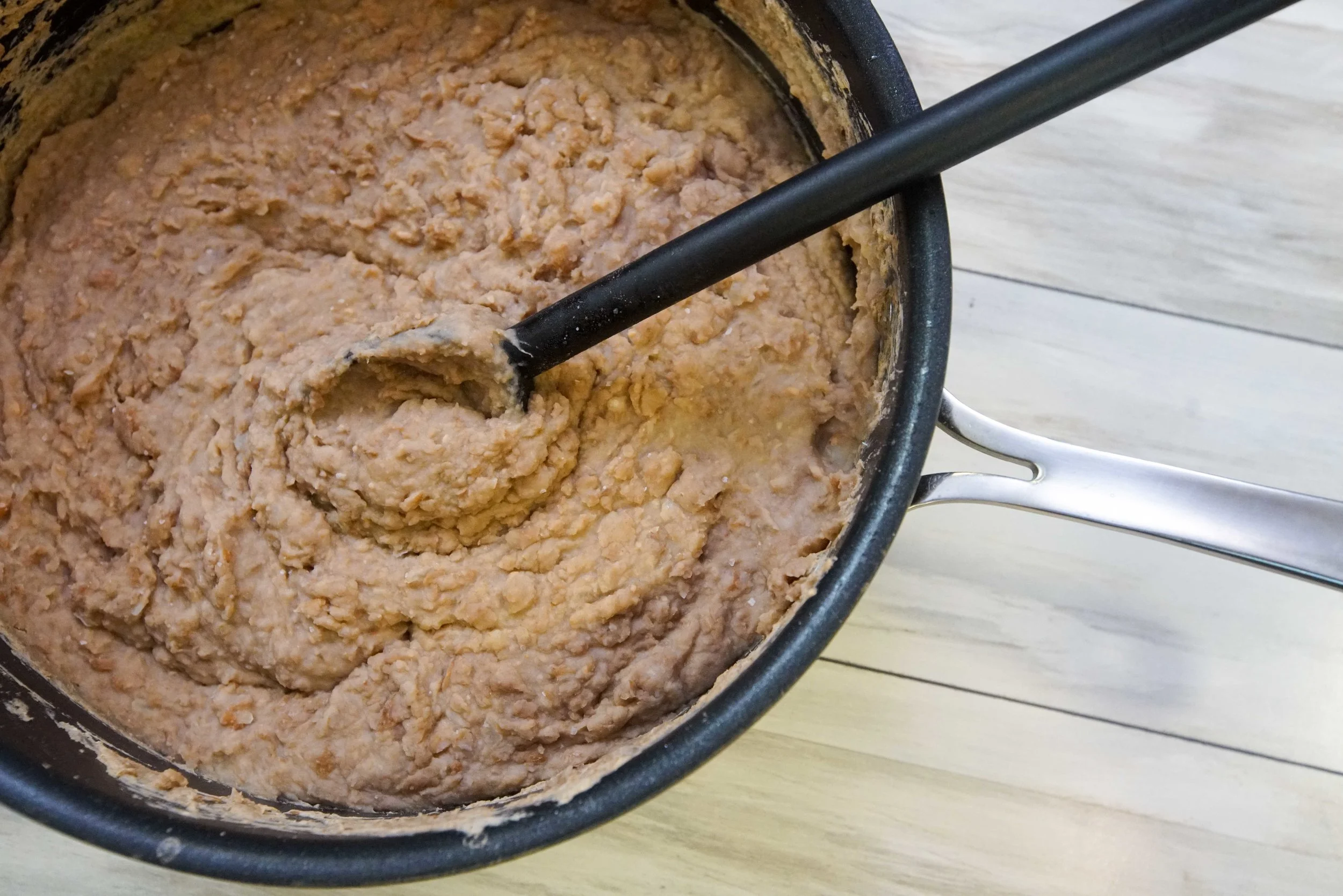 A real photograph of creamy refried beans being stirred in a small saucepan on a stovetop with a wooden spoon