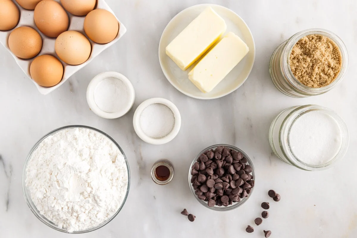 A real photograph of measured flour, brown sugar, granulated sugar, butter, eggs, vanilla, baking soda, salt, and chocolate chips arranged on a tidy kitchen counter with mixing bowls