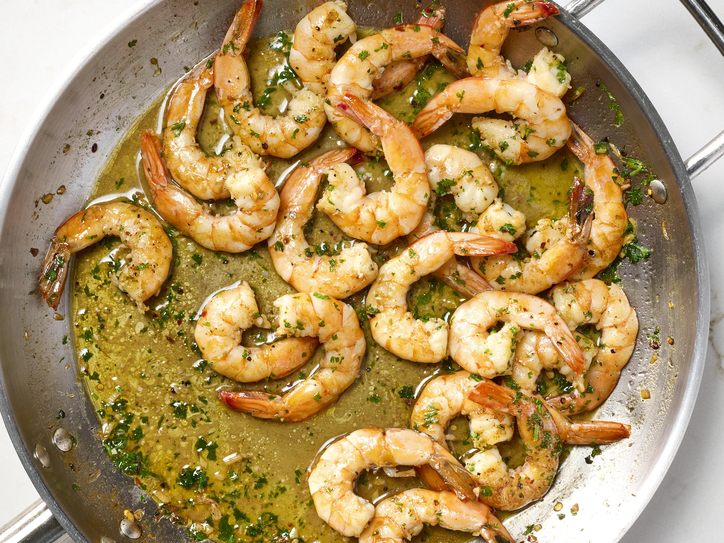 A real photograph of shrimp scampi in a stainless steel skillet, with pink shrimp in a buttery garlic lemon sauce, chopped parsley on top, and a lemon wedge on the side, warm kitchen lighting