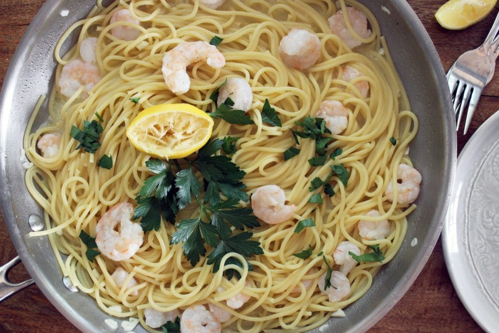 A real photograph of shrimp scampi in a stainless steel skillet with linguine, glossy garlic butter sauce, lemon wedges, and chopped parsley on top