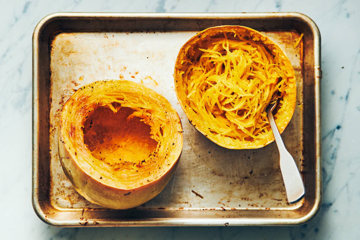 A real photograph of spaghetti squash strands being scraped with a fork inside a roasted squash half on a baking sheet