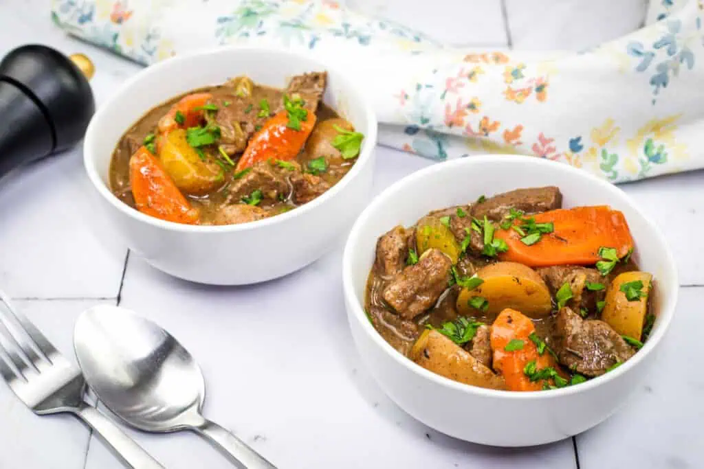 A real photograph of two bowls of beef stew on a table with a spoon and a simple napkin, showing tender beef, carrots, and potatoes in rich gravy