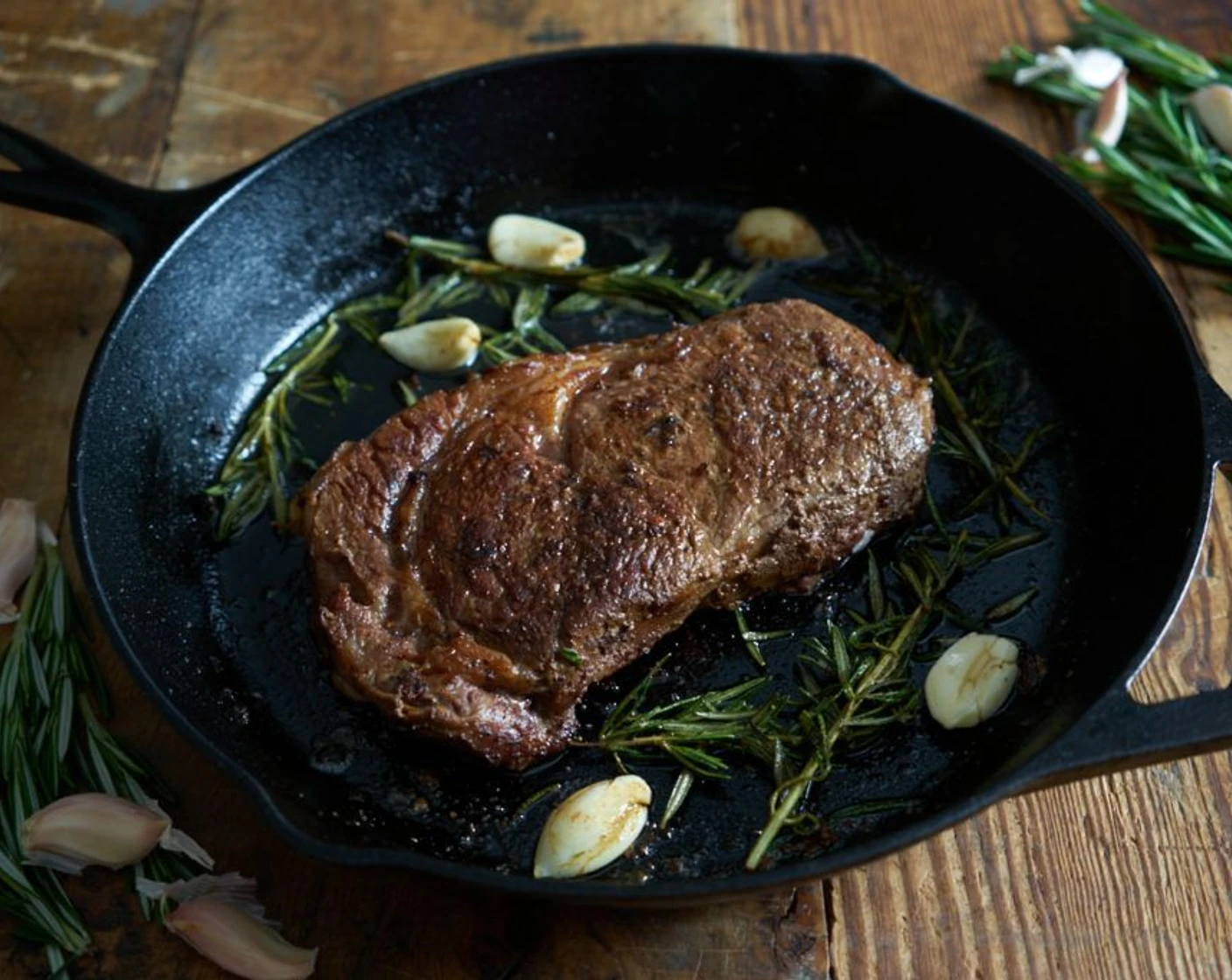 A ribeye steak sizzling in a cast iron skillet with foaming butter, garlic cloves, and thyme on a stovetop