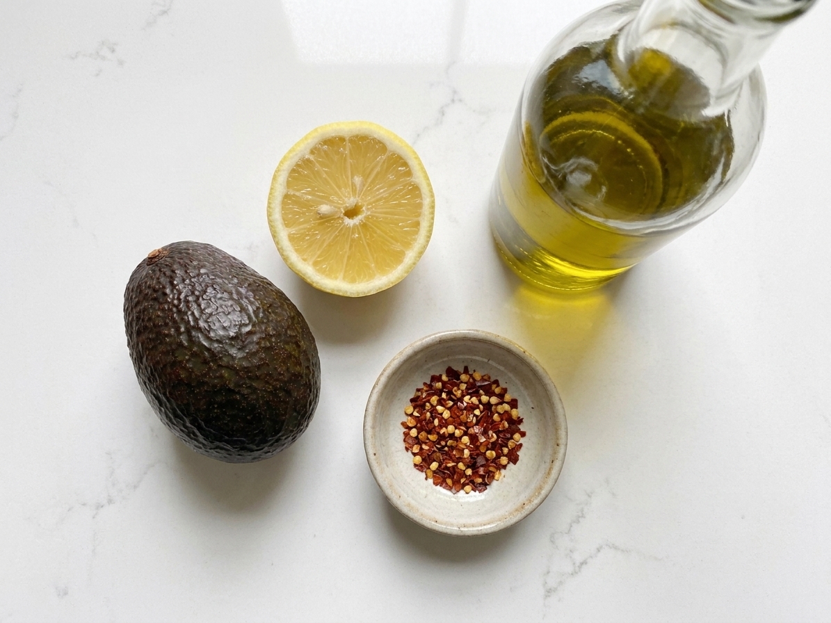 A ripe avocado, a halved lemon, a small dish of red pepper flakes, and a bottle of olive oil arranged on a clean kitchen counter