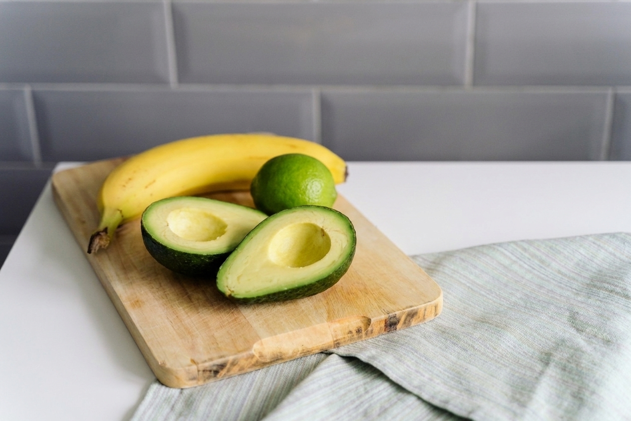A ripe avocado cut in half with the pit removed on a cutting board beside a banana and a lime on a bright kitchen counter