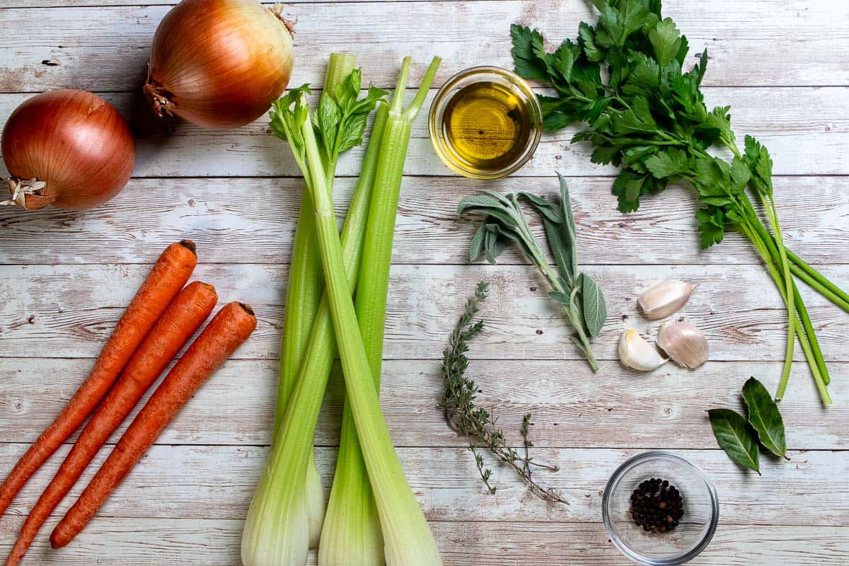 A roasted turkey carcass on a cutting board surrounded by halved onions, carrots, celery stalks, and a small bowl of peppercorns on a home kitchen counter