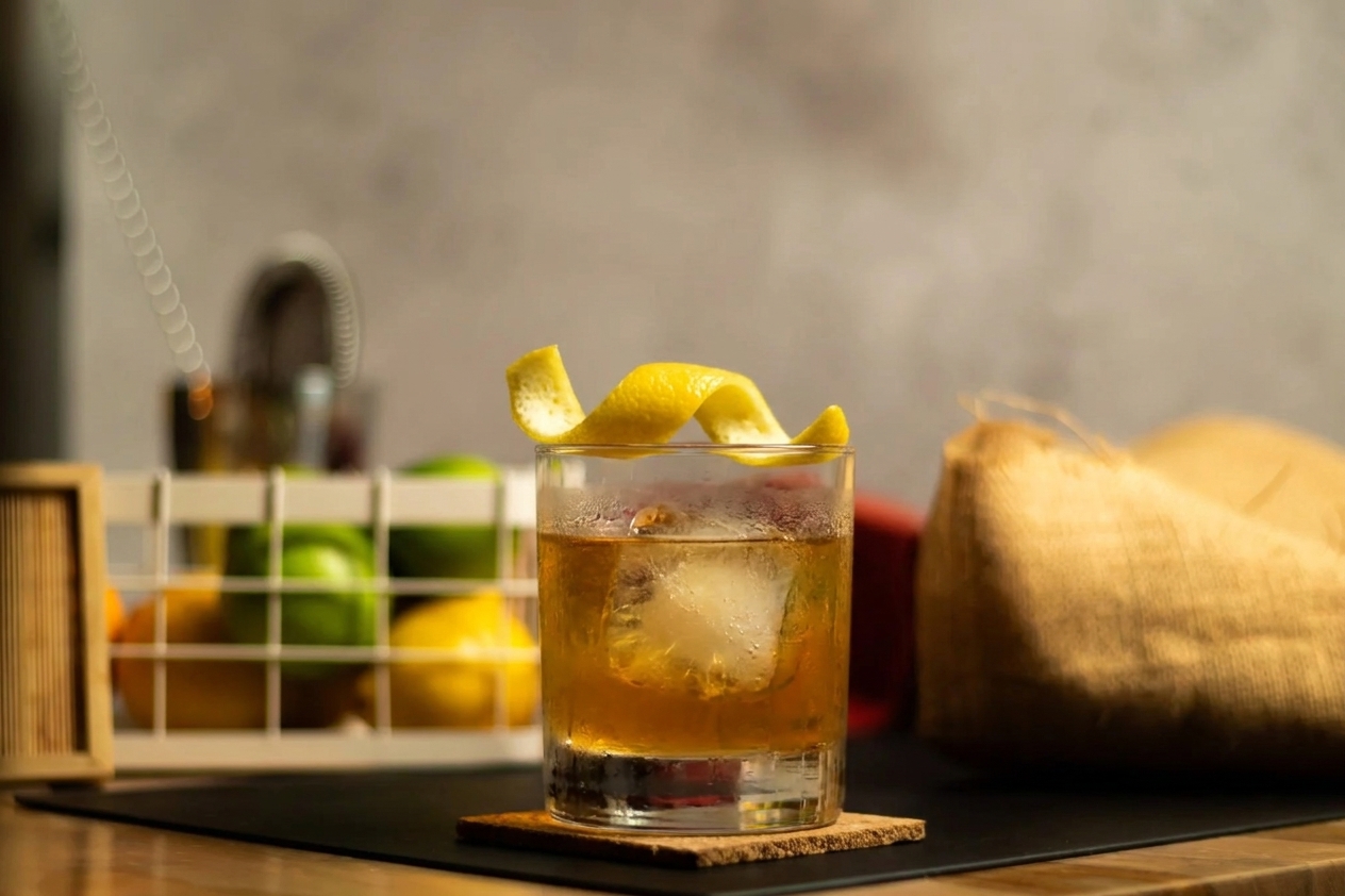 A rocks glass cocktail with a lemon twist garnish resting on a large ice cube, showing condensation on the glass