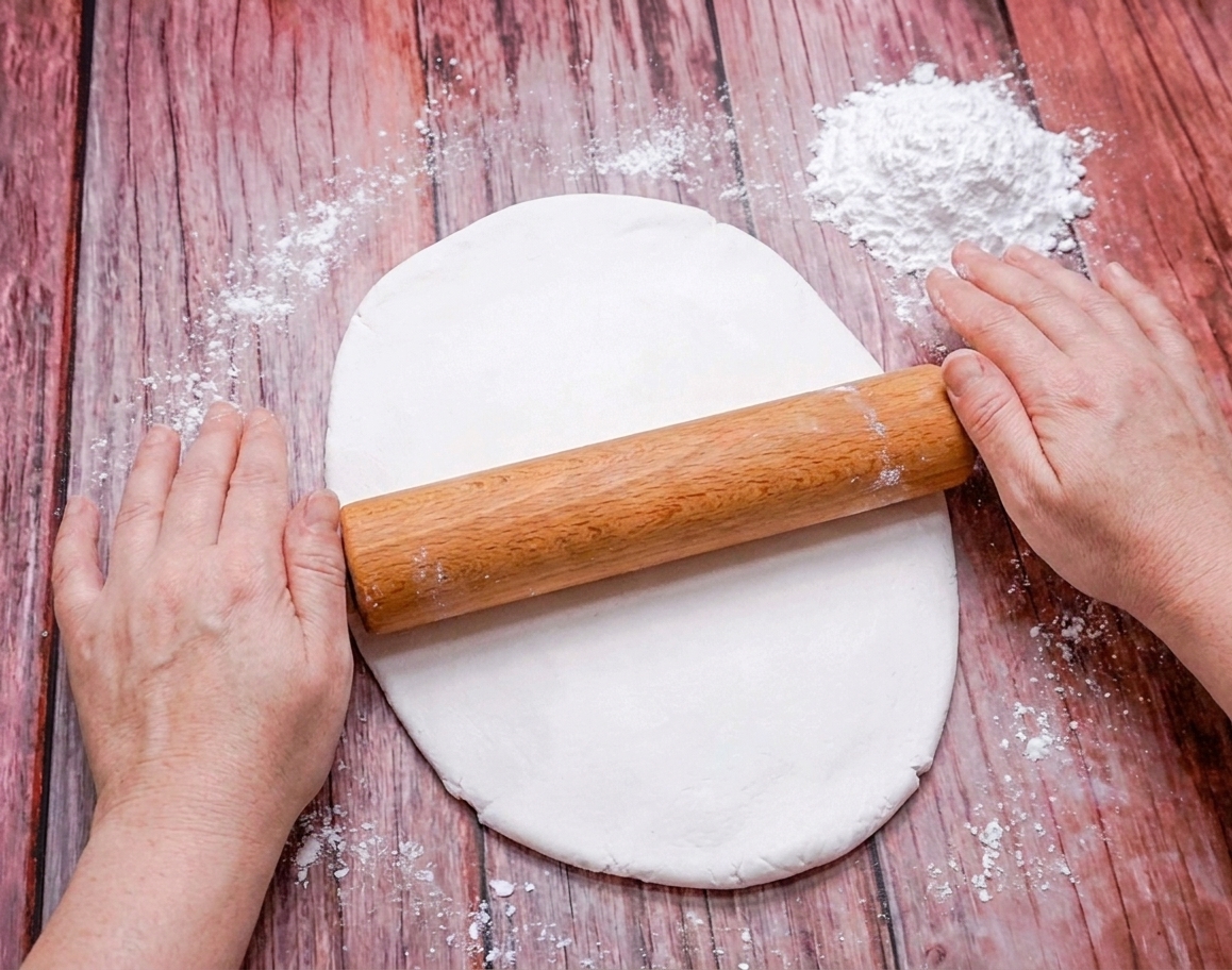 A rolling pin rolling out a sheet of white fondant on a lightly greased counter with powdered sugar nearby