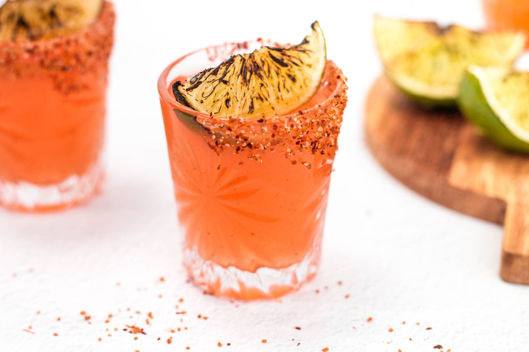 A row of Mexican candy shots with Tajín-chile rims on a warm kitchen counter, small lime wedges and a sliced jalapeño nearby, soft natural light
