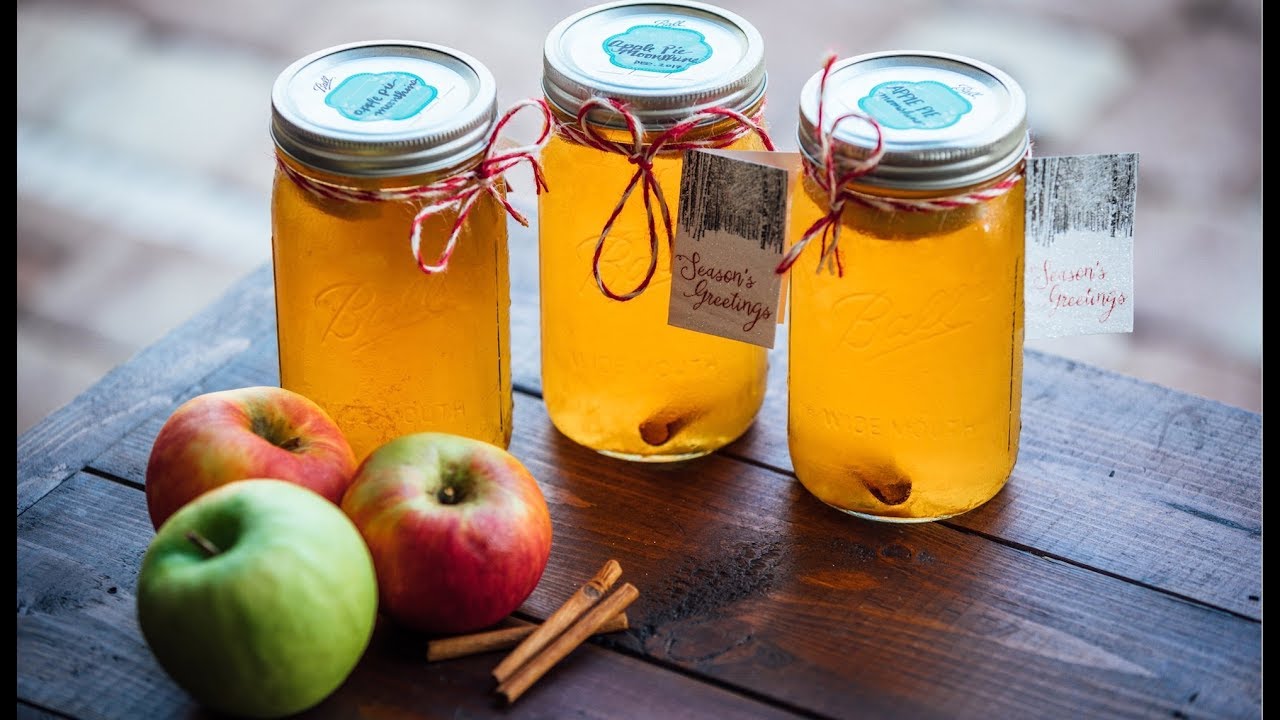 A row of mason jars filled with amber apple cobbler moonshine on a wooden kitchen counter, with cinnamon sticks and sliced apples nearby