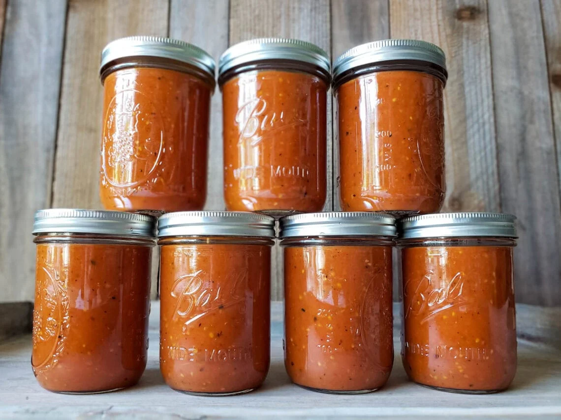 A row of sealed glass mason jars filled with deep red tomato sauce cooling on a wooden kitchen counter with a canning pot nearby