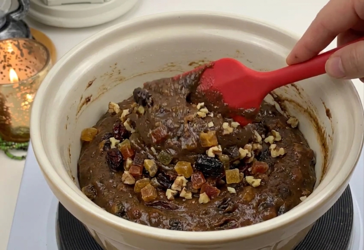 A rubber spatula folding dried fruit and chopped nuts into thick cake batter in a large mixing bowl