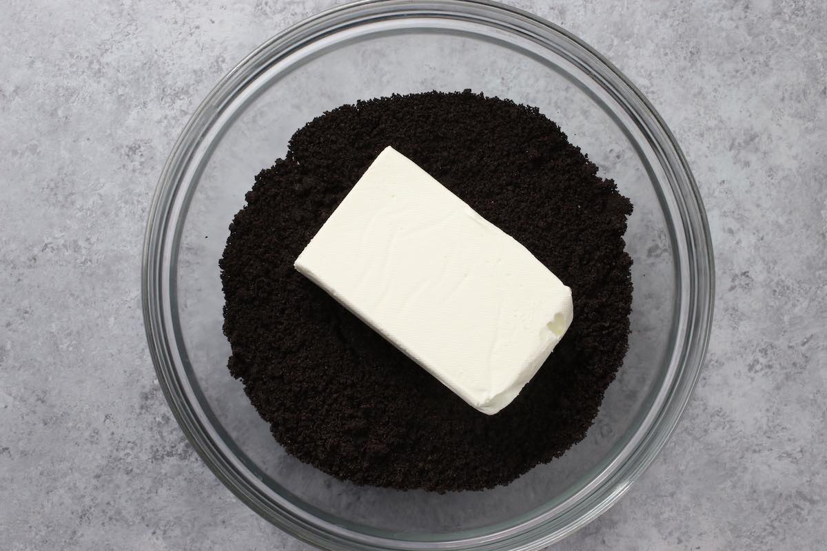 A rubber spatula mixing Oreo cookie crumbs with softened cream cheese in a glass bowl until a thick dough forms on a kitchen counter