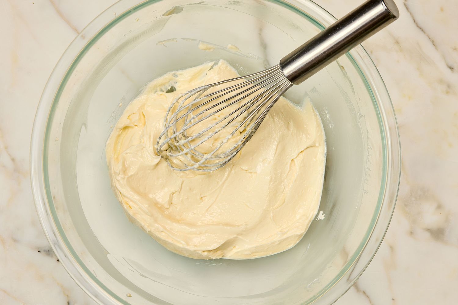 A rubber spatula mixing softened cream cheese and mayonnaise in a white mixing bowl on a kitchen counter