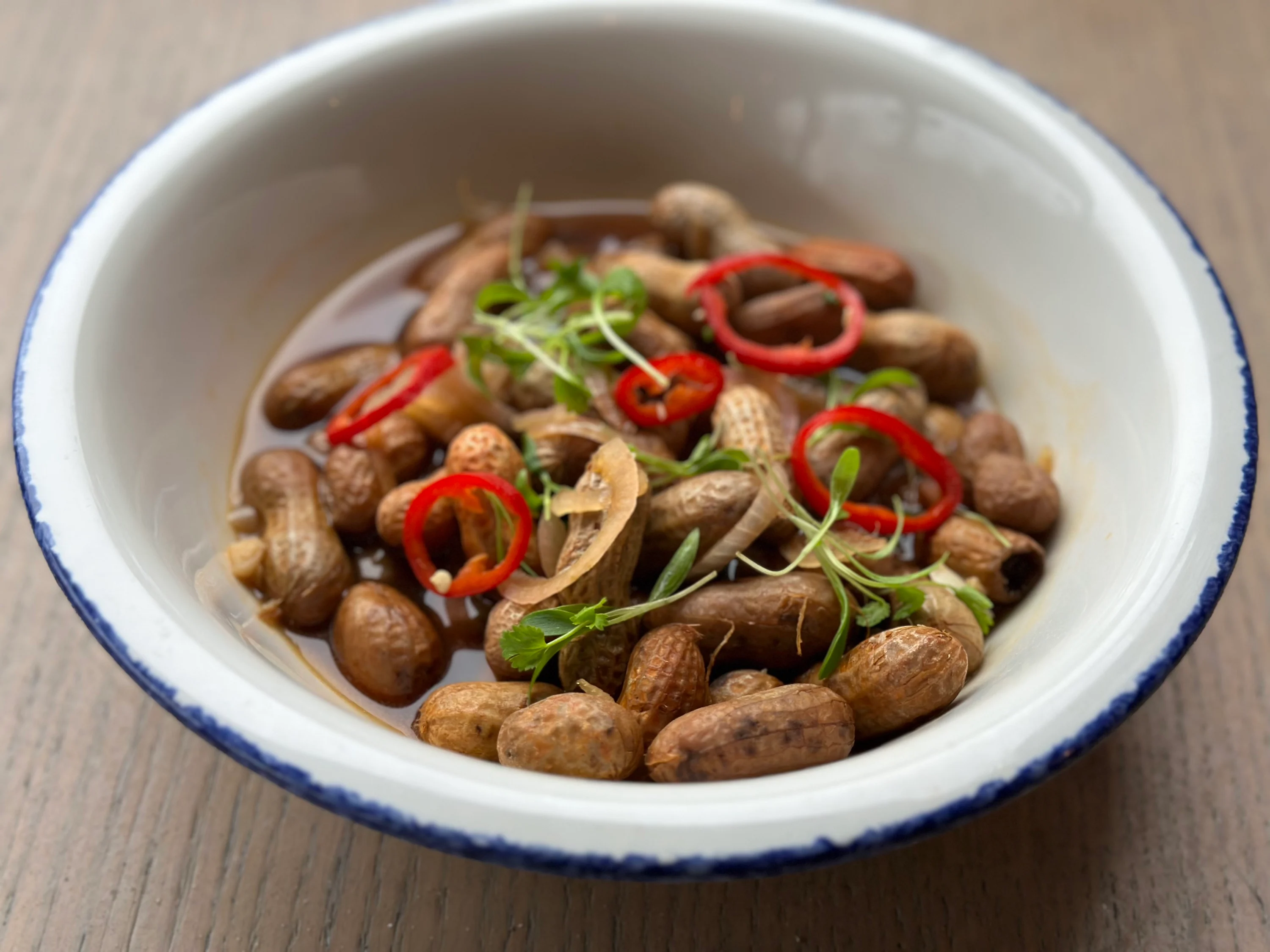 A rustic bowl filled with freshly boiled groundnuts (boiled peanuts) with shells on, lightly wet and glossy, set on a wooden kitchen table with a linen towel